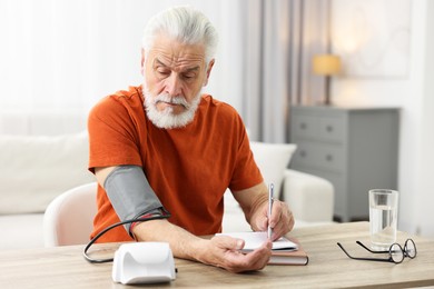 Senior man writing results of blood pressure measurement at table indoors