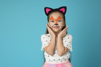 Cute girl with painted face and ears as cat on light blue background, space for text