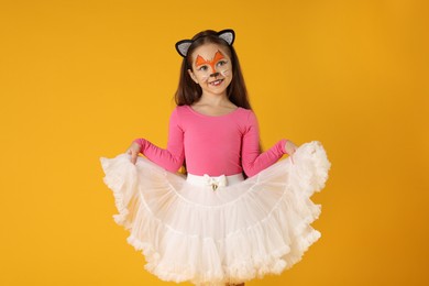 Cute girl with painted face and ears as cat on orange background