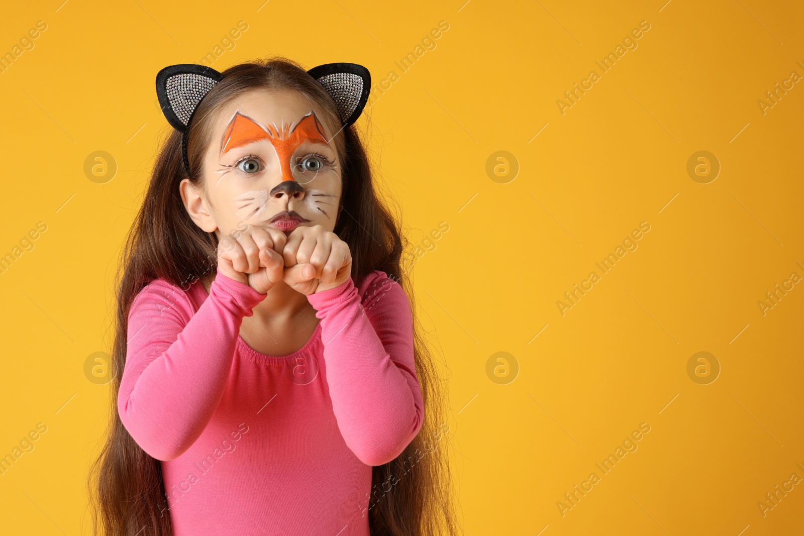 Photo of Cute girl with painted face and ears as cat on orange background, space for text