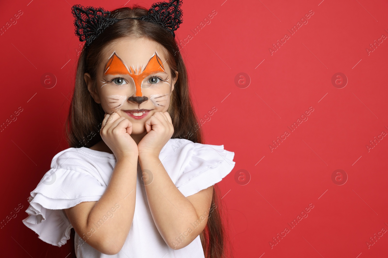 Photo of Cute girl with painted face and ears as cat on red background, space for text