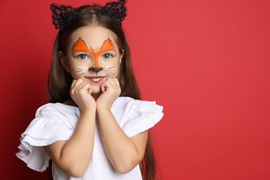 Photo of Cute girl with painted face and ears as cat on red background, space for text