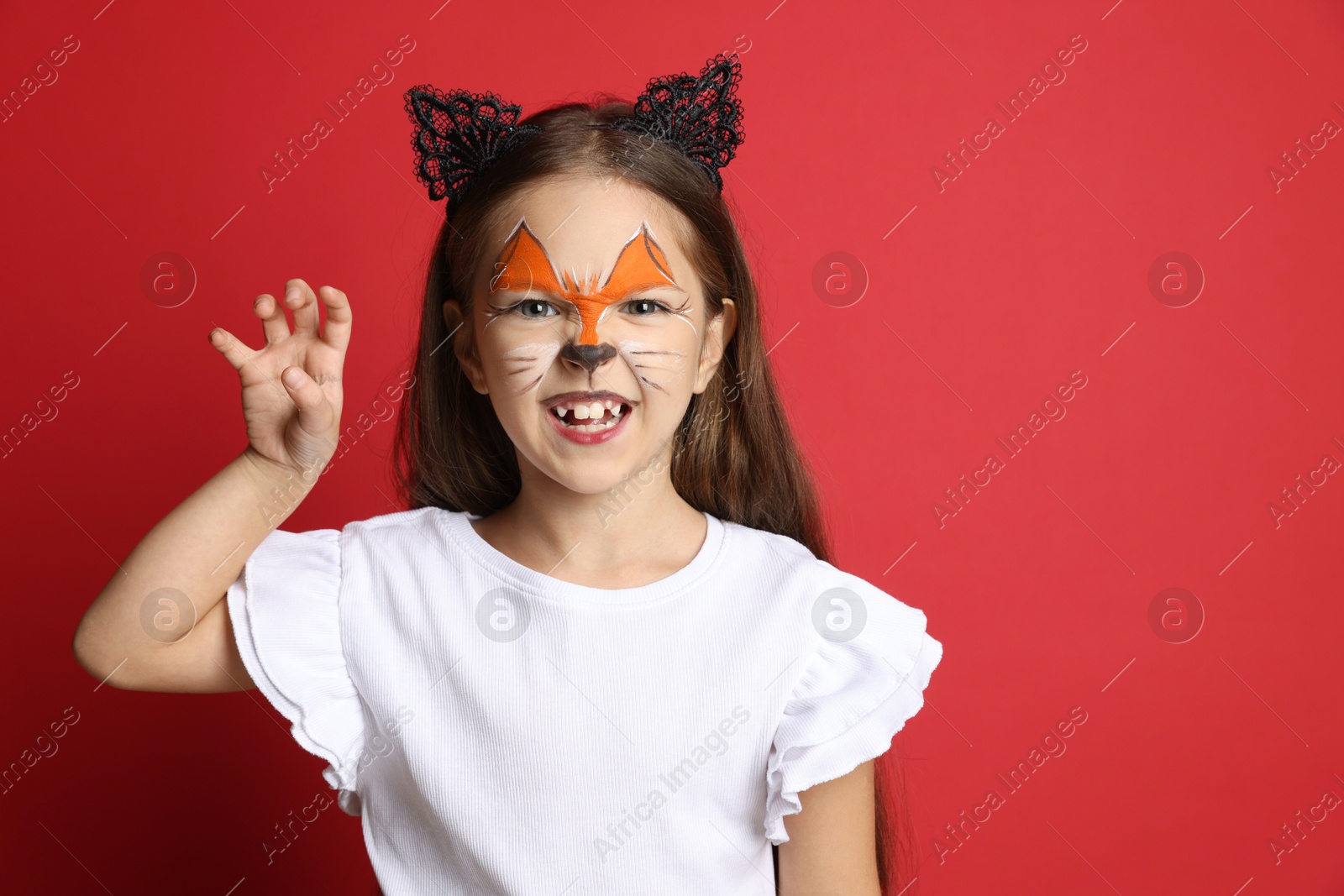 Photo of Cute girl with painted face and ears as cat on red background, space for text