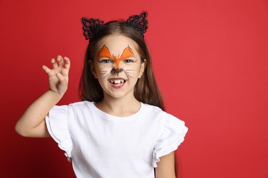 Photo of Cute girl with painted face and ears as cat on red background, space for text