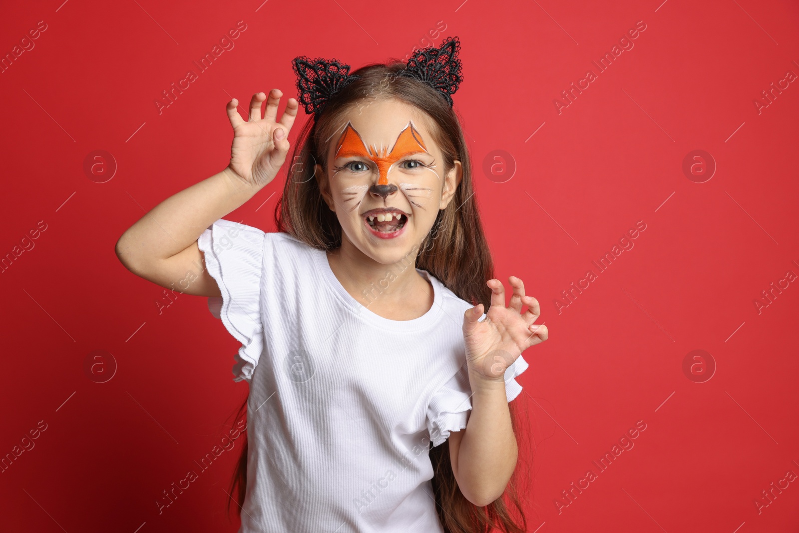 Photo of Cute girl with painted face and ears as cat on red background, space for text