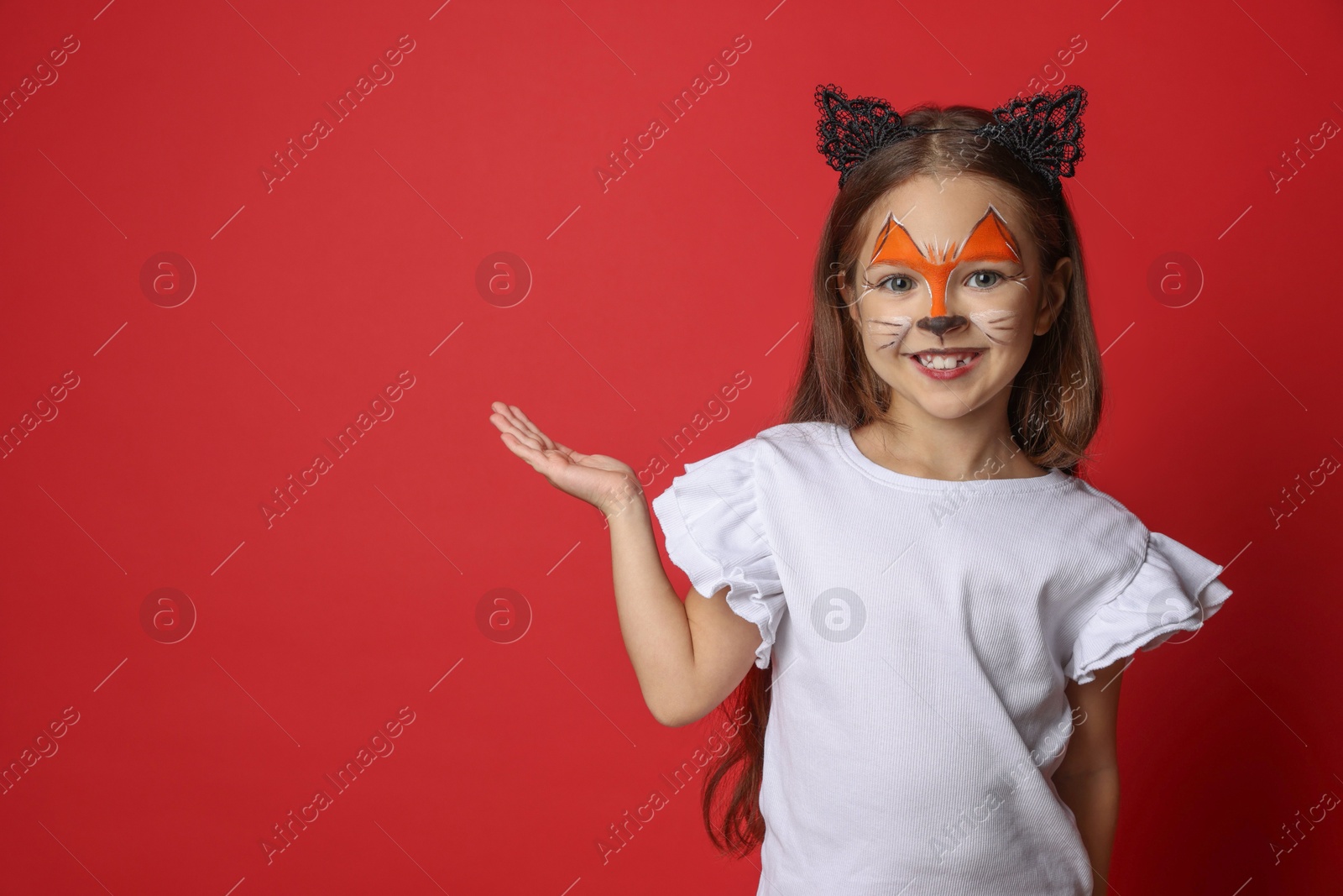 Photo of Cute girl with painted face and ears as cat on red background, space for text