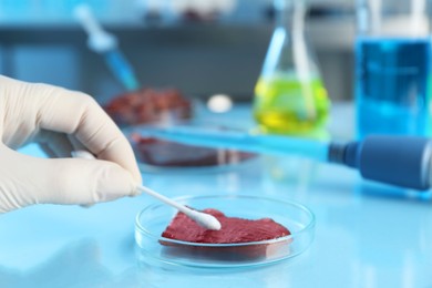 Photo of GMO concept. Scientist with swab and piece of meat at table in laboratory, closeup. Space for text