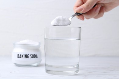 Photo of Woman adding baking soda to glass of water at white textured table, closeup