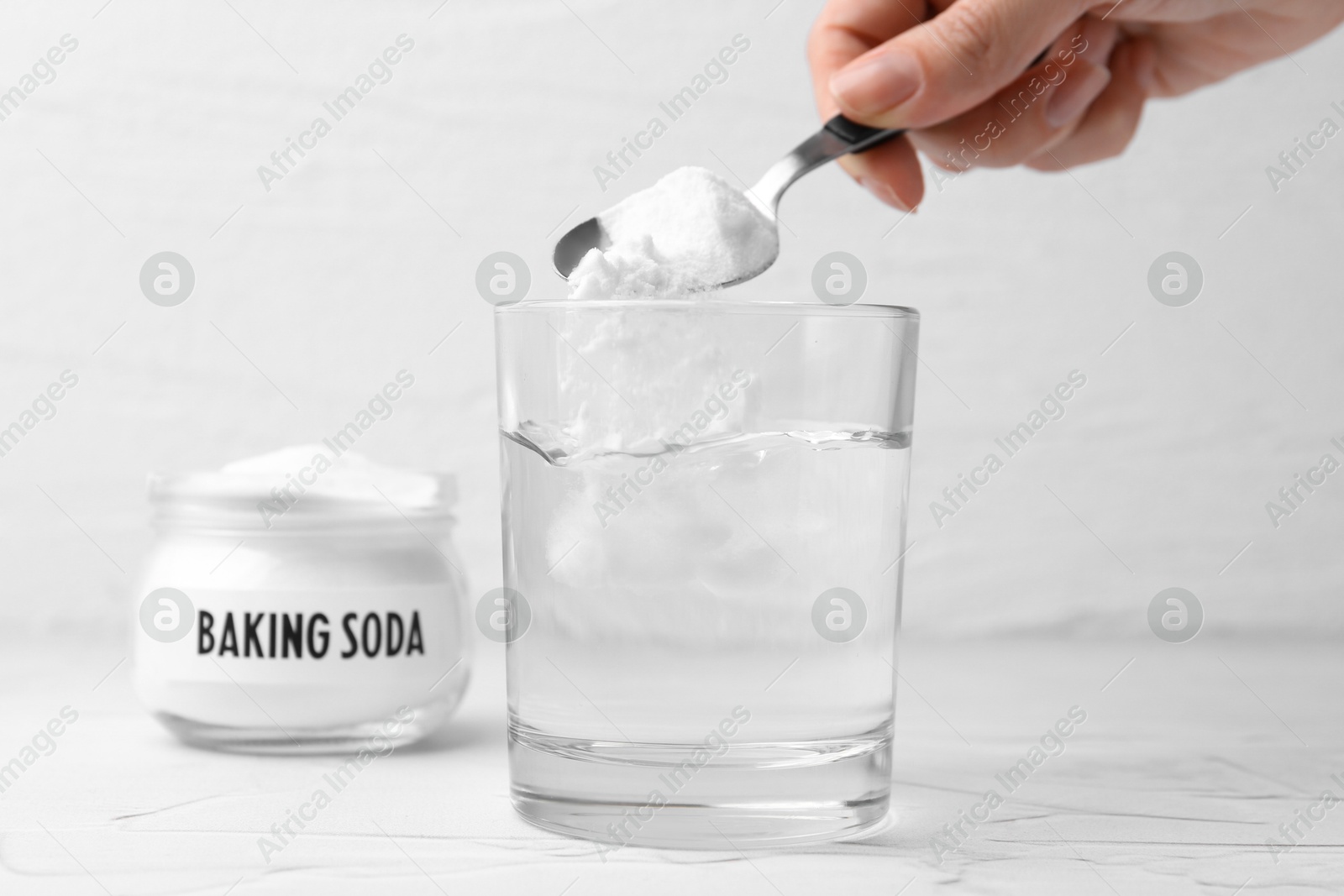 Photo of Woman adding baking soda to glass of water at white textured table, closeup