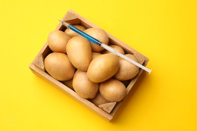 Photo of GMO concept. Potatoes and syringe with liquid in crate on yellow background, top view