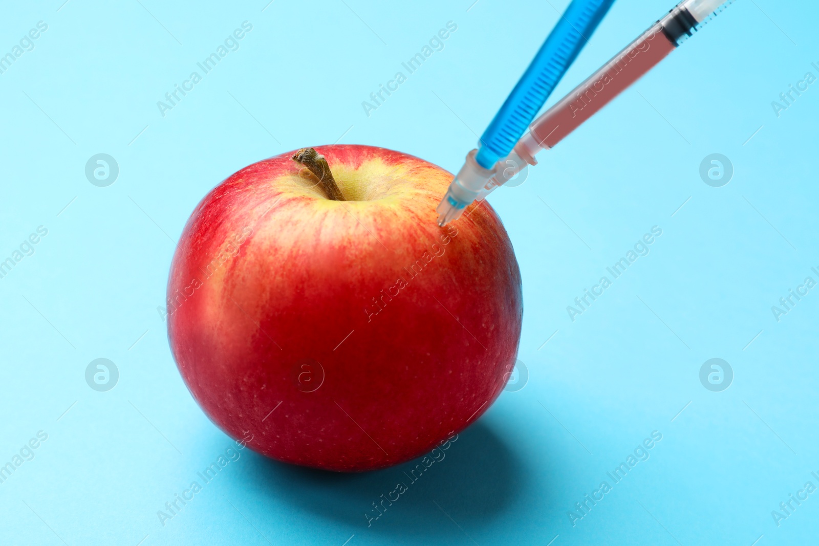 Photo of GMO concept. Red apple with different syringes on light blue background, closeup