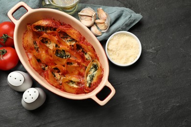 Photo of Delicious conchiglie pasta with ricotta cheese, spinach and tomato sauce in baking dish on black textured table, flat lay. Space for text