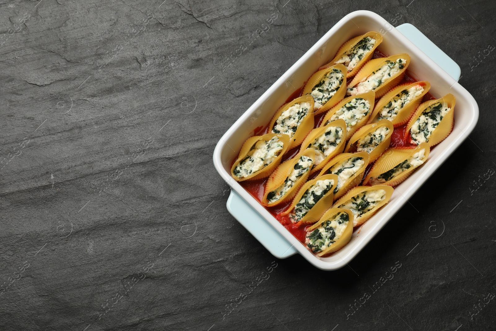 Photo of Delicious conchiglie pasta with ricotta cheese, spinach and tomato sauce in baking dish on black textured table, top view. Space for text