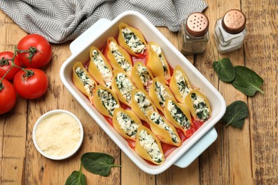 Photo of Delicious conchiglie pasta with ricotta cheese, spinach and tomato sauce in baking dish on wooden table, flat lay