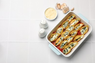 Photo of Delicious conchiglie pasta with ricotta cheese, spinach and tomato sauce in baking dish on white tiled table, flat lay. Space for text