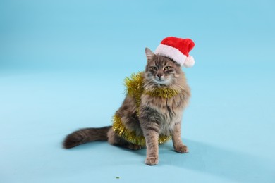 Photo of Cute cat in Santa hat with yellow Christmas tinsel on light blue background