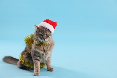 Photo of Cute cat in Santa hat with yellow Christmas tinsel on light blue background, space for text