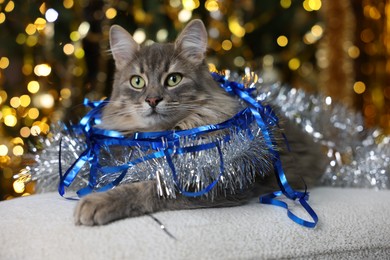 Photo of Cute cat with shiny tinsels on pouf against blurred lights. Christmas atmosphere