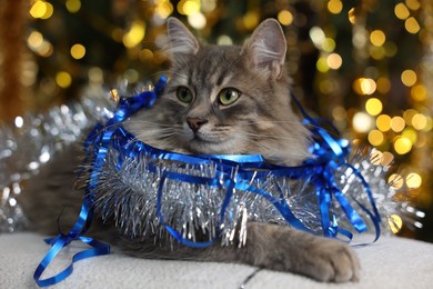 Photo of Cute cat with shiny tinsels on pouf against blurred lights. Christmas atmosphere