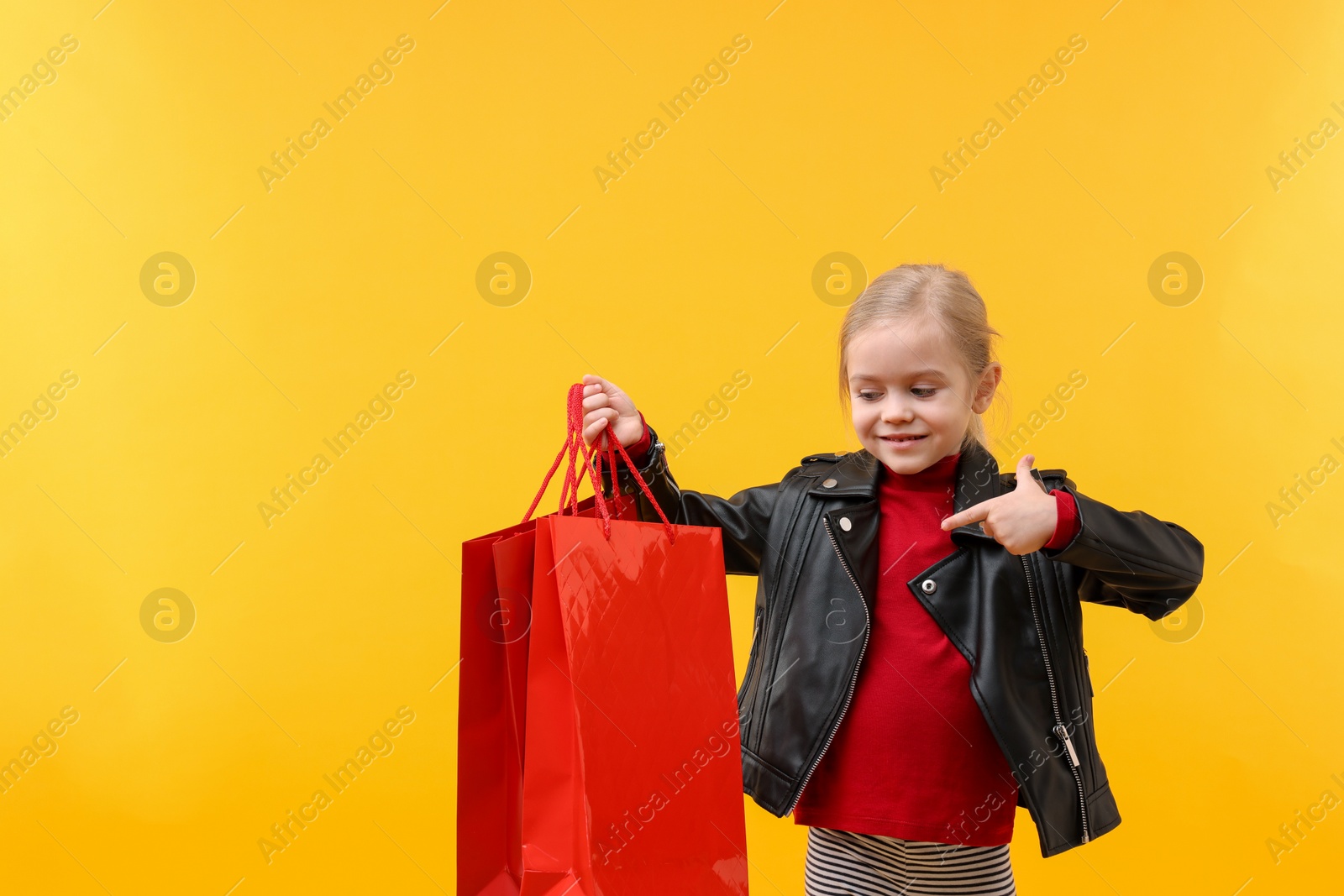 Photo of Little girl pointing at shopping bags on orange background, space for text