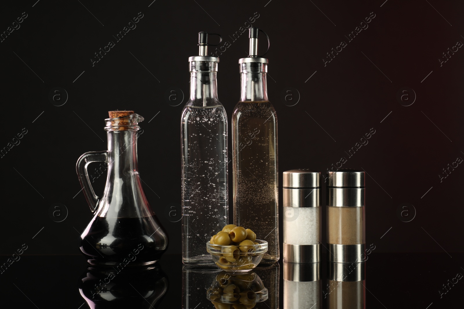 Photo of Salad dressings, spices and olives on mirror surface against dark background