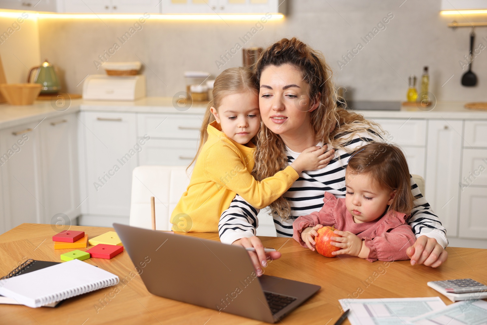 Photo of Single mother working on laptop and her daughters at table in kitchen