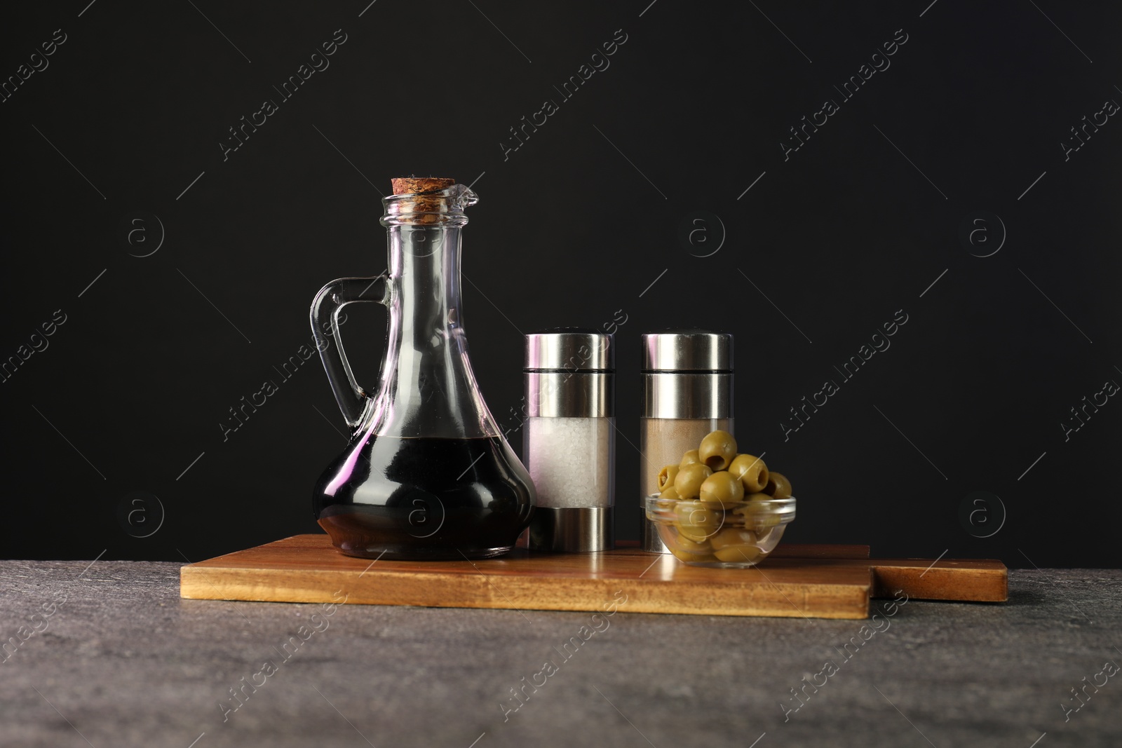 Photo of Vinegar in bottle, olives and spices on grey table against black background