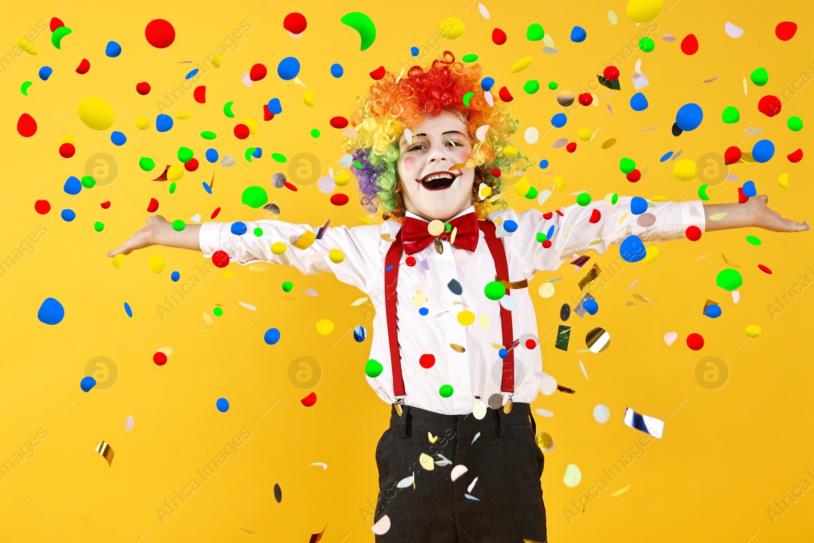 Image of Happy boy dressed like clown under falling confetti on orange background. Party time