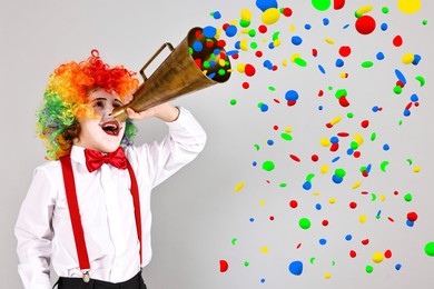 Image of Cute boy dressed like clown shouting in megaphone with confetti flying out of it on grey background. Party time