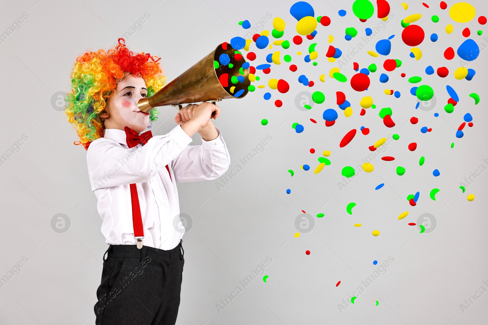 Image of Cute boy dressed like clown shouting in megaphone with confetti flying out of it on grey background. Party time