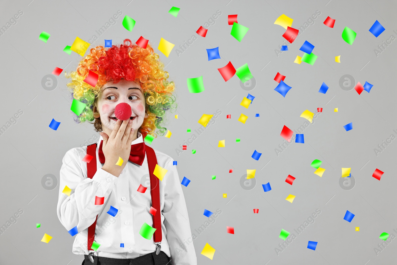 Image of Cute boy dressed like clown under falling confetti on grey background. Party time