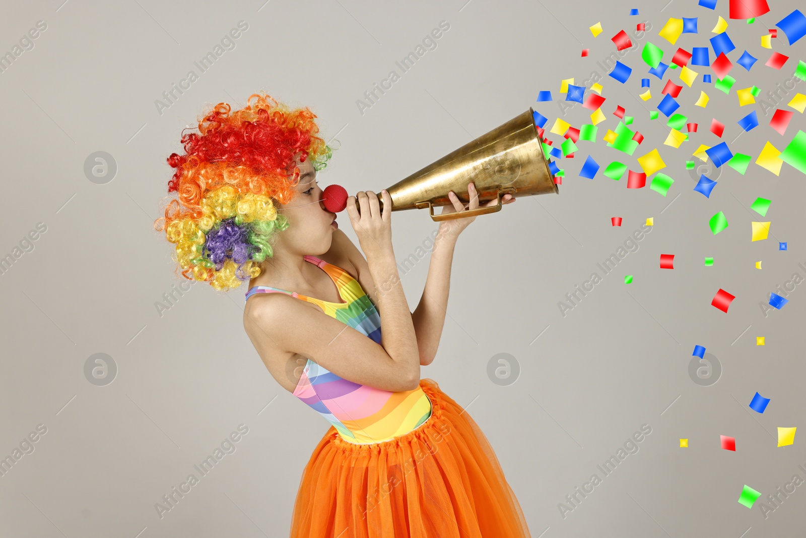 Image of Cute girl dressed like clown shouting in megaphone with confetti flying out of it on grey background. Party time