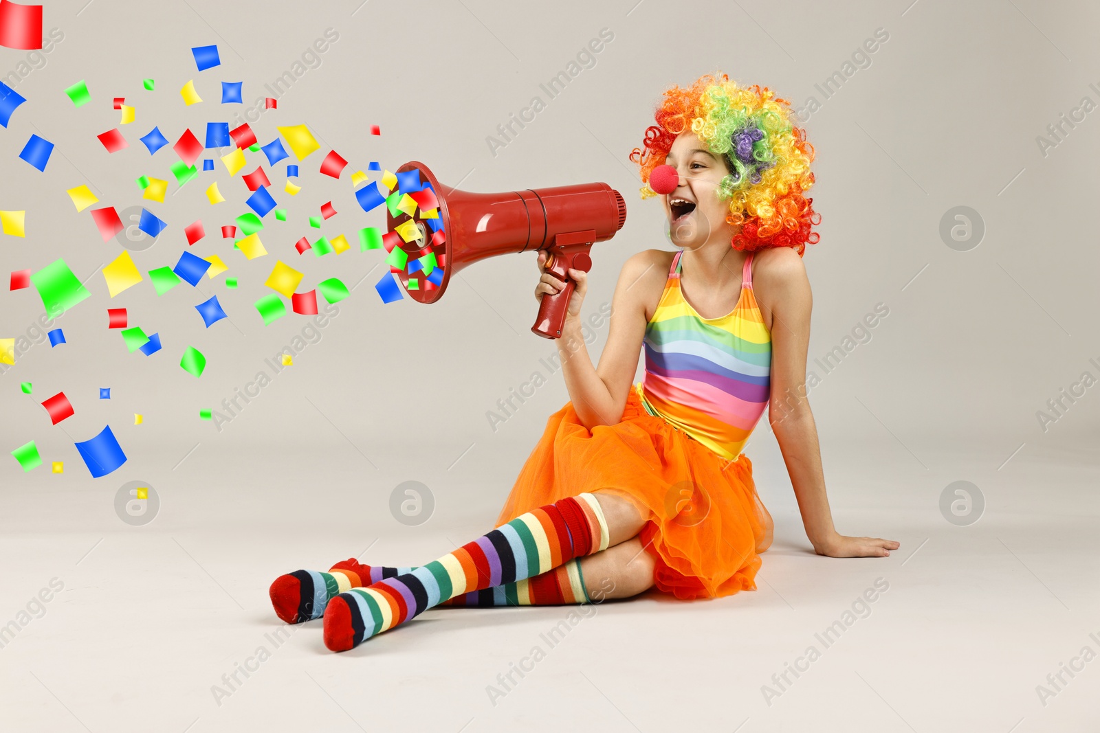 Image of Cute girl dressed like clown shouting in megaphone with confetti flying out of it on grey background. Party time