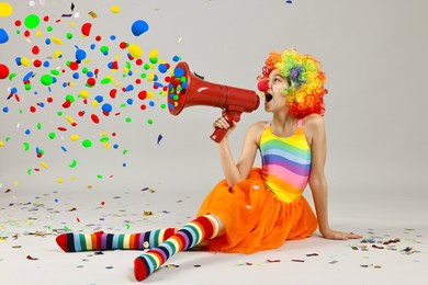 Image of Cute girl dressed like clown shouting in megaphone with confetti flying out of it on grey background. Party time