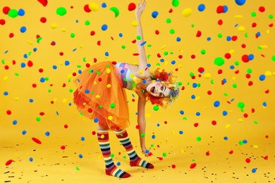 Happy girl dressed like clown under falling confetti on orange background. Party time