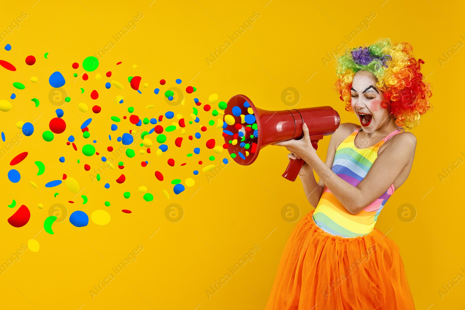 Image of Cute girl dressed like clown shouting in megaphone with confetti flying out of it on orange background. Party time