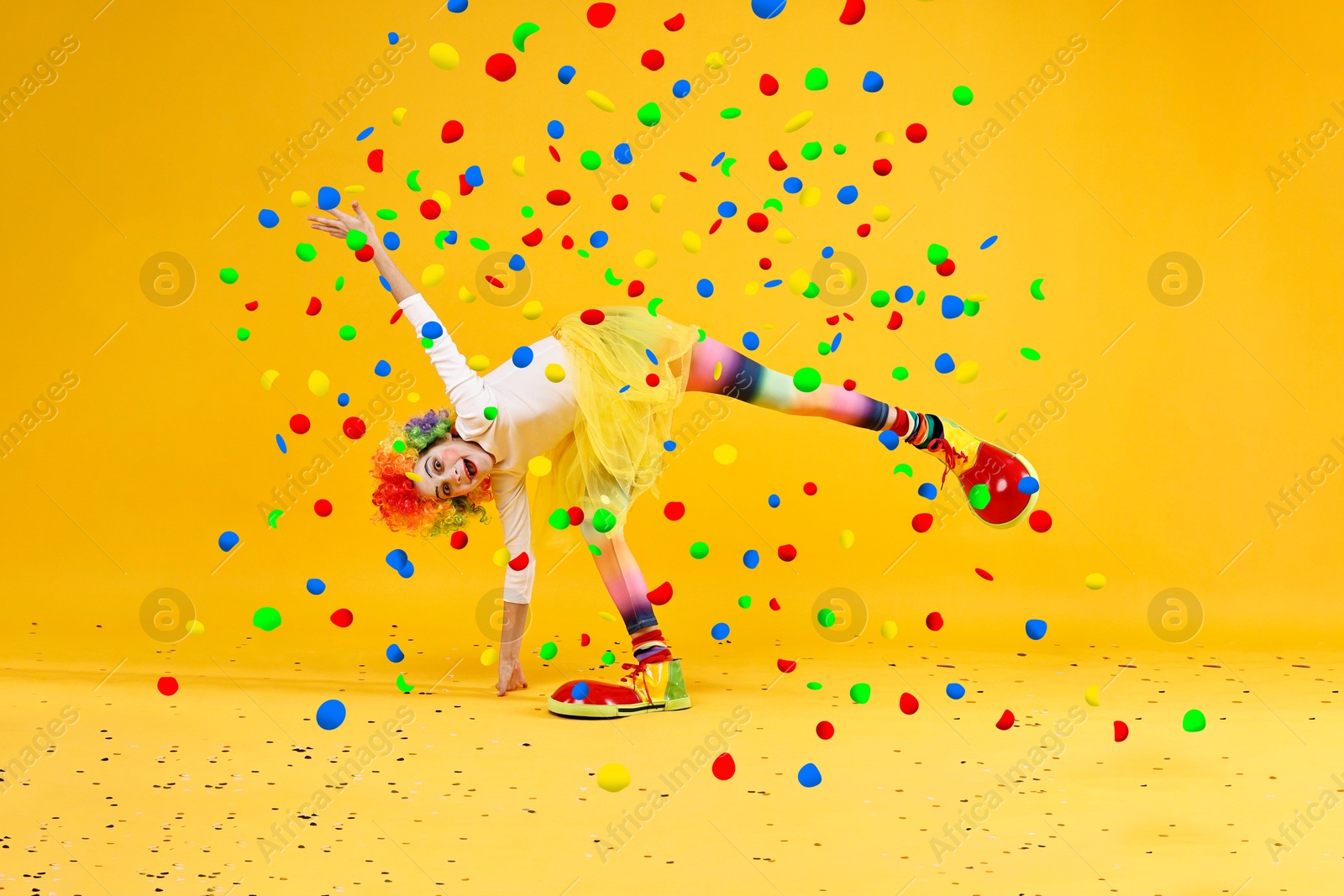 Image of Happy girl dressed like clown under falling confetti on orange background. Party time