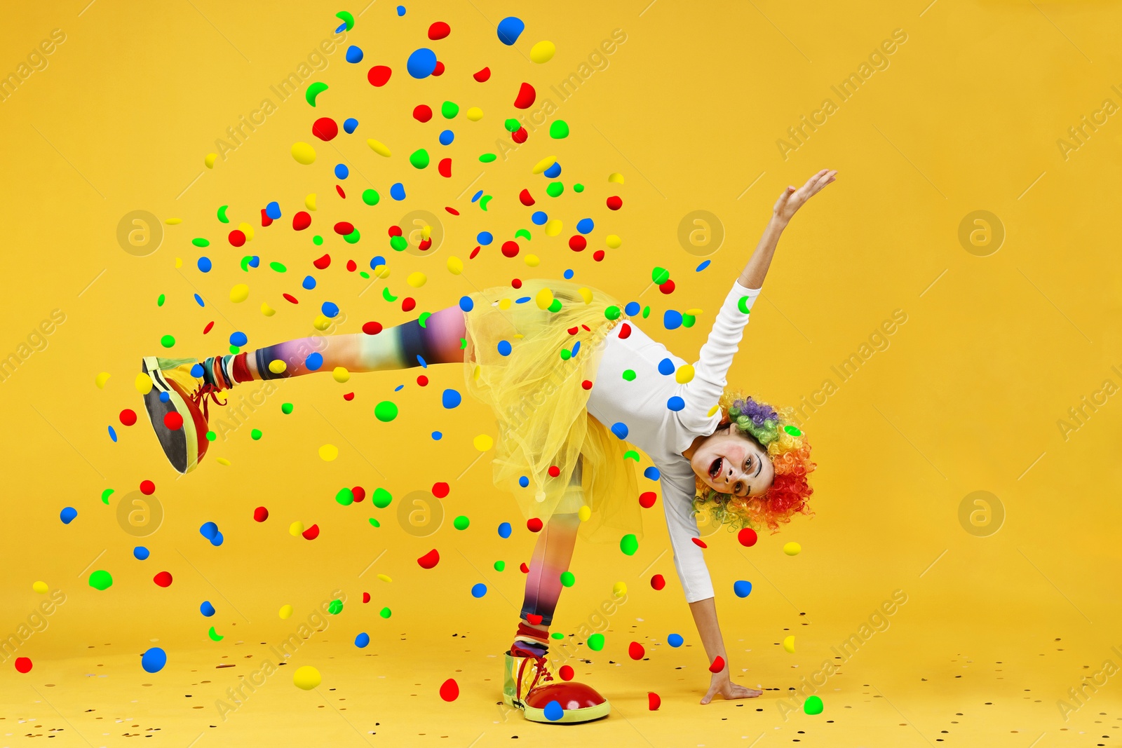 Image of Happy girl dressed like clown under falling confetti on orange background. Party time