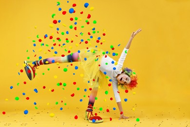 Happy girl dressed like clown under falling confetti on orange background. Party time