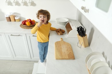 Photo of Little boy eating cookie while standing on step stool near countertop in kitchen