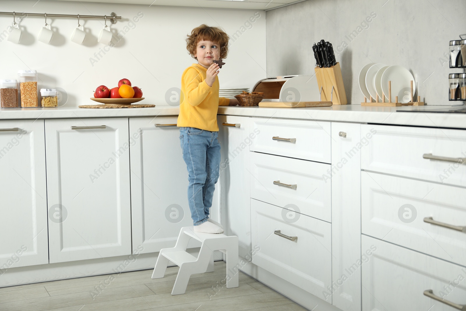 Photo of Little boy with cookie standing on step stool in kitchen