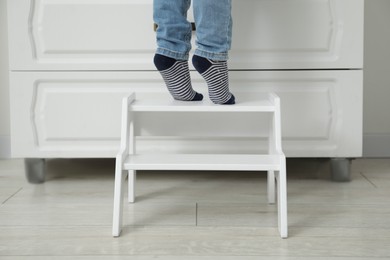 Photo of Little boy standing on step stool near chest of drawers at home, closeup