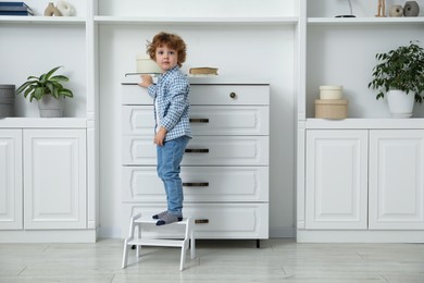 Photo of Little boy standing on step stool near chest of drawers at home