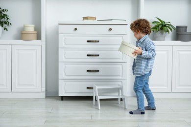 Photo of Little boy with box and step stool near chest of drawers at home