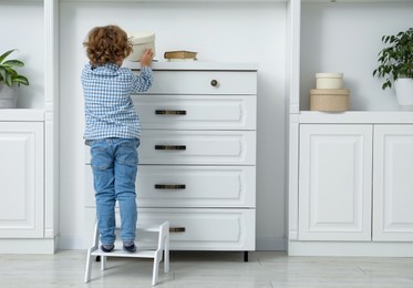 Photo of Little boy standing on step stool and reaching for box at home, back view