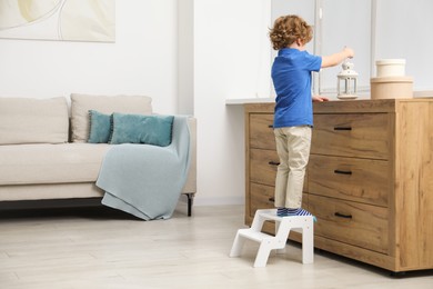 Photo of Little boy with lantern standing on step stool near chest of drawers at home