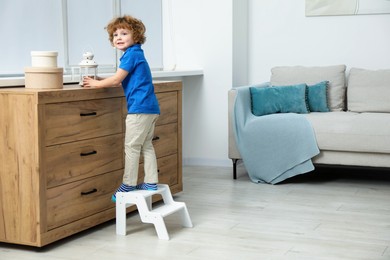 Photo of Little boy with lantern standing on step stool near chest of drawers at home