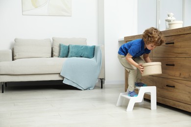 Photo of Little boy with box standing on step stool near chest of drawers at home