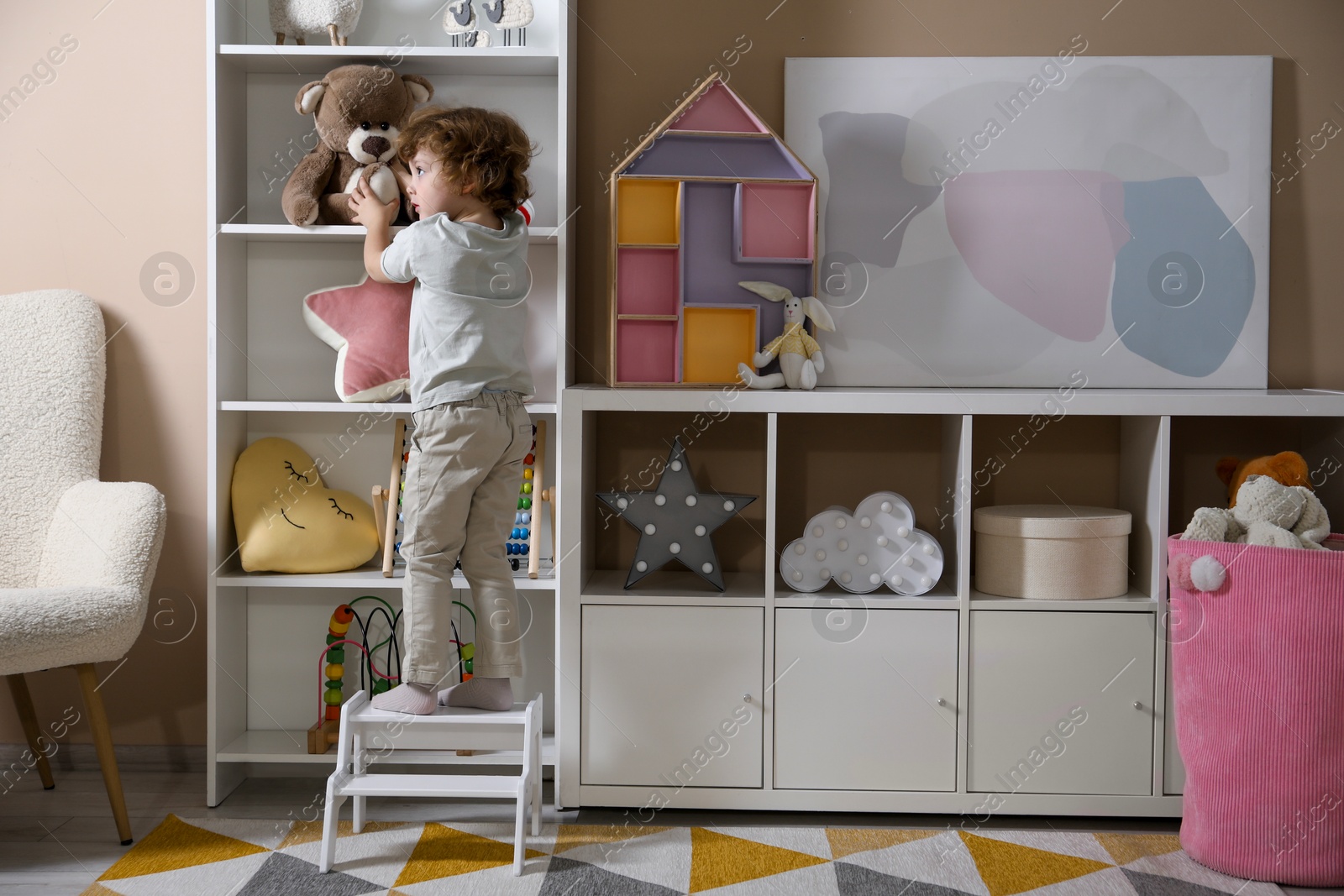 Photo of Little boy standing on step stool and reaching for toys at home