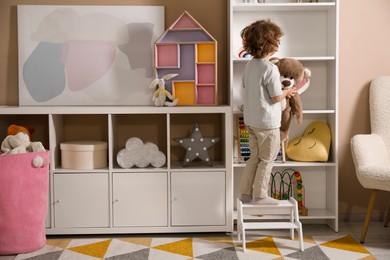 Photo of Little boy with teddy bear standing on step stool at home